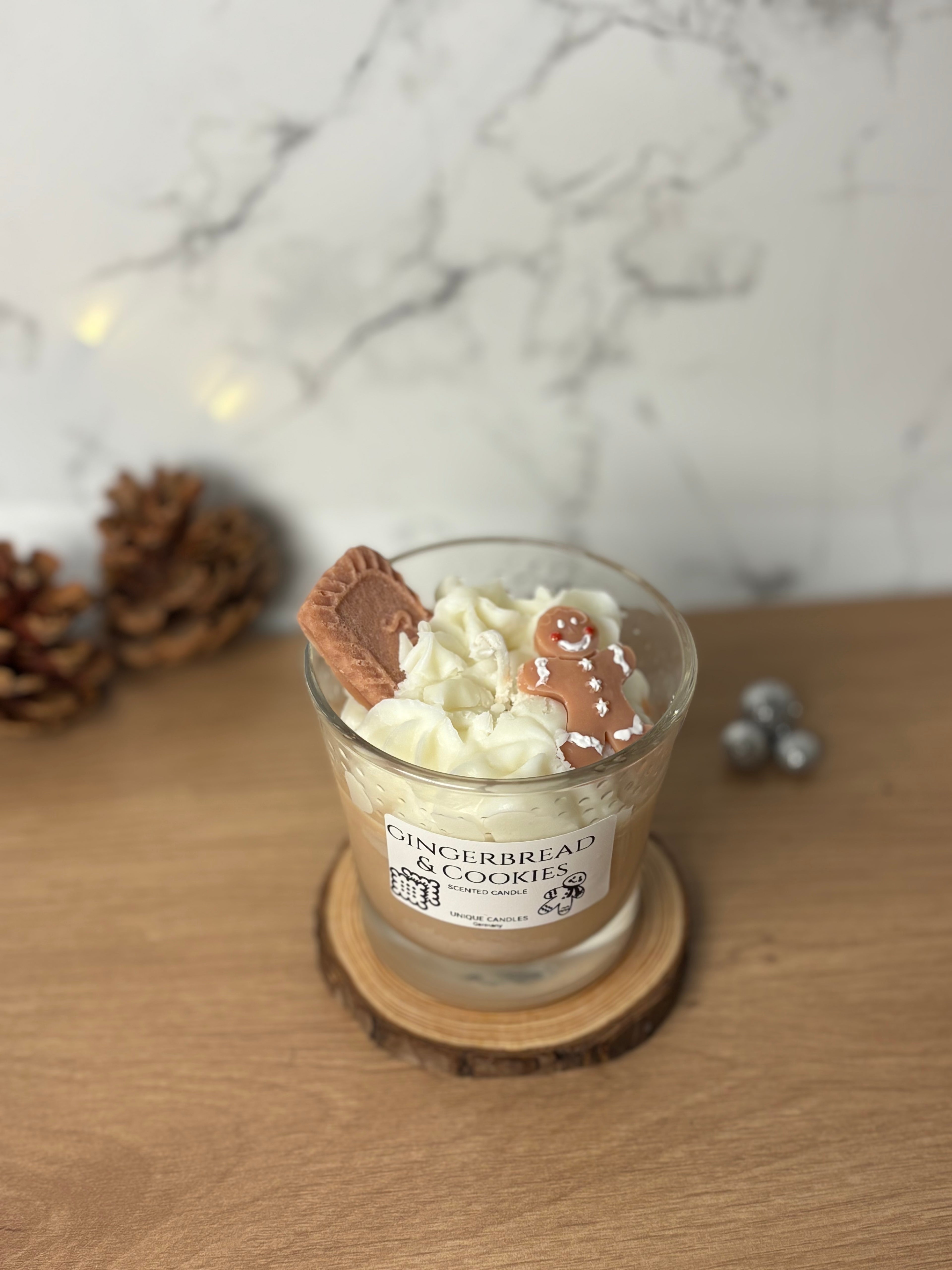 Handgegossene Bio-Sojawachs Duftkerze "Gingerbread & Cookies" im Glas auf einem Holzuntersetzer. Die Kerze hat eine cremige Wachs-Sahnehaube, dekoriert mit einem kleinen Spekulatius-Keks und einem Lebkuchenmann aus Wachs. Perfekte Weihnachtsdeko mit Spekulatiusduft.