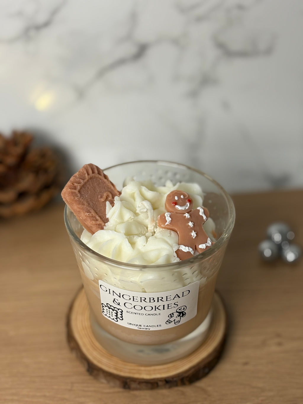 Handgegossene Bio-Sojawachs Duftkerze "Gingerbread & Cookies" im Glas auf einem Holzuntersetzer. Die Kerze hat eine cremige Wachs-Sahnehaube, dekoriert mit einem kleinen Spekulatius-Keks und einem Lebkuchenmann aus Wachs. Perfekte Weihnachtsdeko mit Spekulatiusduft.